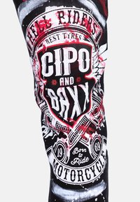 Pantalon en tissu noir avec des imprimés graphiques blancs et rouges de motifs liés à la moto, contenant du texte, le tout avec une texture usée.