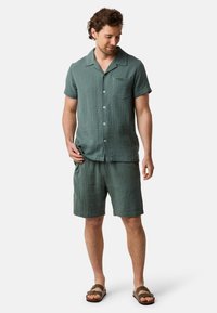 Homme portant une chemise à manches courtes verte à boutons et un short assorti, se tenant les mains détendues, regardant vers le bas et souriant, sur fond blanc.