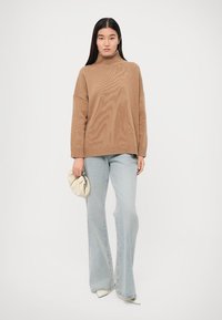 Pull à col roulé beige avec texture côtelée, coupe oversize, associé à un jean évasé bleu clair et un sac à main de couleur claire. Des talons blancs complètent l'ensemble.