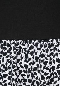 Musta paita, jossa on teksturoitu, istuva yläosa ja levenevä alaosa, joka koostuu valkoisesta kankaasta mustilla lehtikuvioilla, kokoontuneena saumaan.