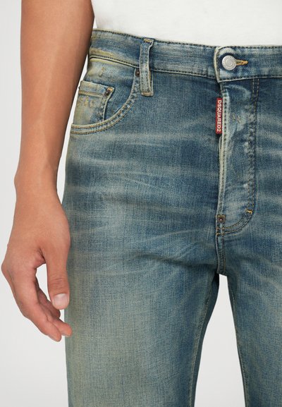 Jeans in denim blu con un lavaggio sbiadito, lievi segni di usura, design a cinque tasche, chiusura con bottone metallico e etichetta rossa in vita.