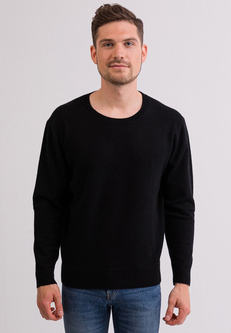 Schwarzer Strickpullover mit rundem Ausschnitt, langen Ärmeln und gerippten Bündchen. Getragen mit blauen Jeans, vor einem hellen Hintergrund präsentiert.