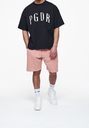 Schwarzes Baumwoll-T-Shirt mit weißem "PGDR"-Aufdruck, kombiniert mit rosa Textur-Shorts. Weiße Sneakers und schwarz-weiße Socken vervollständigen das Outfit.