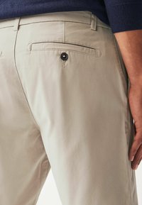 Pantalones beiges hechos de una tela suave, con un bolsillo trasero que tiene un botón negro y un ajuste entallado. Se puede ver costura acentuada.
