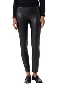 Schwarze enganliegende Leggings aus Kunstleder mit hoher Taille, vertikalen Nähten und einer glatten, glänzenden Textur. Getragen mit schwarzen Slippern.