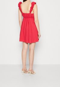 Robe rouge sans manches avec bretelles volantes, taille élastique, jupe évasée et texture lisse. Portée avec des sandales à talons de couleur claire.