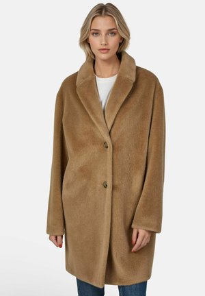 Vrouw die een oversized lichtbruine nep-bonte jas met twee knopen draagt over een wit topje en blauwe spijkerbroek, staand tegen een eenvoudige lichte achtergrond.