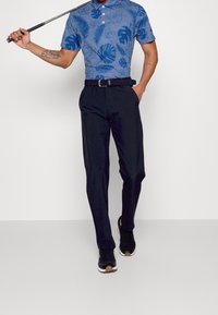 Sininen poolopaita, jossa on lehtikuosisia kuvioita, mustat housut ja mustat tennarit. Paidassa on kaulus ja kolme nappia. Kokonaisuus on suljettu.