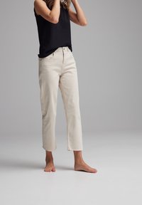 Jeans beige a vita alta con orlo grezzo, abbinati a un top nero senza maniche. Il tessuto appare liscio, con un classico design a cinque tasche.