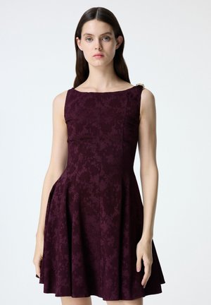 Vrouw met lang donker haar, gekleed in een mouwloze donkerpaarse jurk met een subtiel bloemenpatroon en een getailleerde taille.