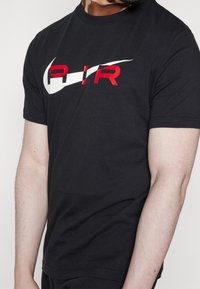 Svart bomullst-shirt med stort, vitt Nike-swoosh och röd "AIR"-text i mitten. Korta ärmar och rund halsdesign.