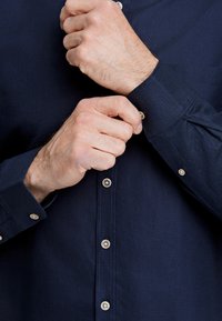 Camicia button-up blu navy con una texture liscia, caratterizzata da bottoni di colore chiaro e polsini regolabili. Cuciture visibili lungo il colletto.