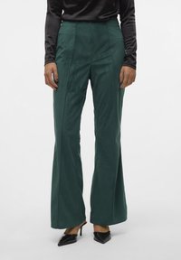 Pantalons évasés en velours côtelé vert avec des côtes verticales, taille haute et poches latérales, associés à des escarpins noires à bout pointu. Texture de tissu lisse.