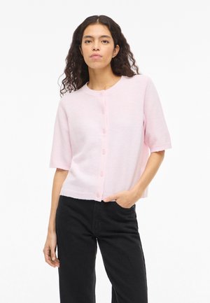 Femme aux cheveux bouclés portant un cardigan à manches courtes rose clair boutonné et un pantalon noir, debout avec une main dans la poche.