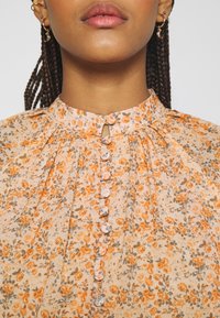Blusa floral en melocotón claro con patrones en naranja y gris. Tiene un escote alto, pliegues y botones cubiertos de tela en la parte delantera.