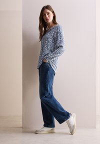 Leichter, langärmliger Oberteil in einem blauen und weißen abstrakten Muster, kombiniert mit dunkelblauen Jeans und weißen Sneakers mit grauen Akzenten.