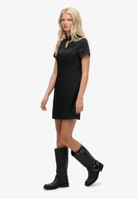 Schwarzes kurzes Kleid mit Kragen und Ausschnittdetail, kombiniert mit kniehohen schwarzen Lederstiefeln mit Schnallen und strukturierter Oberfläche.