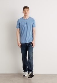 T-shirt de manga curta azul claro com decote redondo, combinado com jeans azul escuro e ténis de cores mistas, exibido contra um fundo neutro.