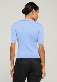 Vrouw met donker haar in een knot, gekleed in een lichtblauw geribd T-shirt met korte mouwen en een zwarte spijkerbroek, staand met haar rug naar de camera tegen een effen achtergrond.