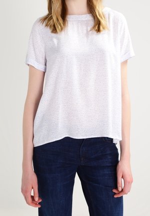 Femme portant un T-shirt blanc texturé à manches courtes et un jean bleu foncé, debout devant un fond neutre.