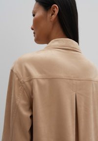 Beige stoffen jacket met een gladde textuur, voorzien van een klassieke kraag en een achterkant met plooidetail voor extra stijl.