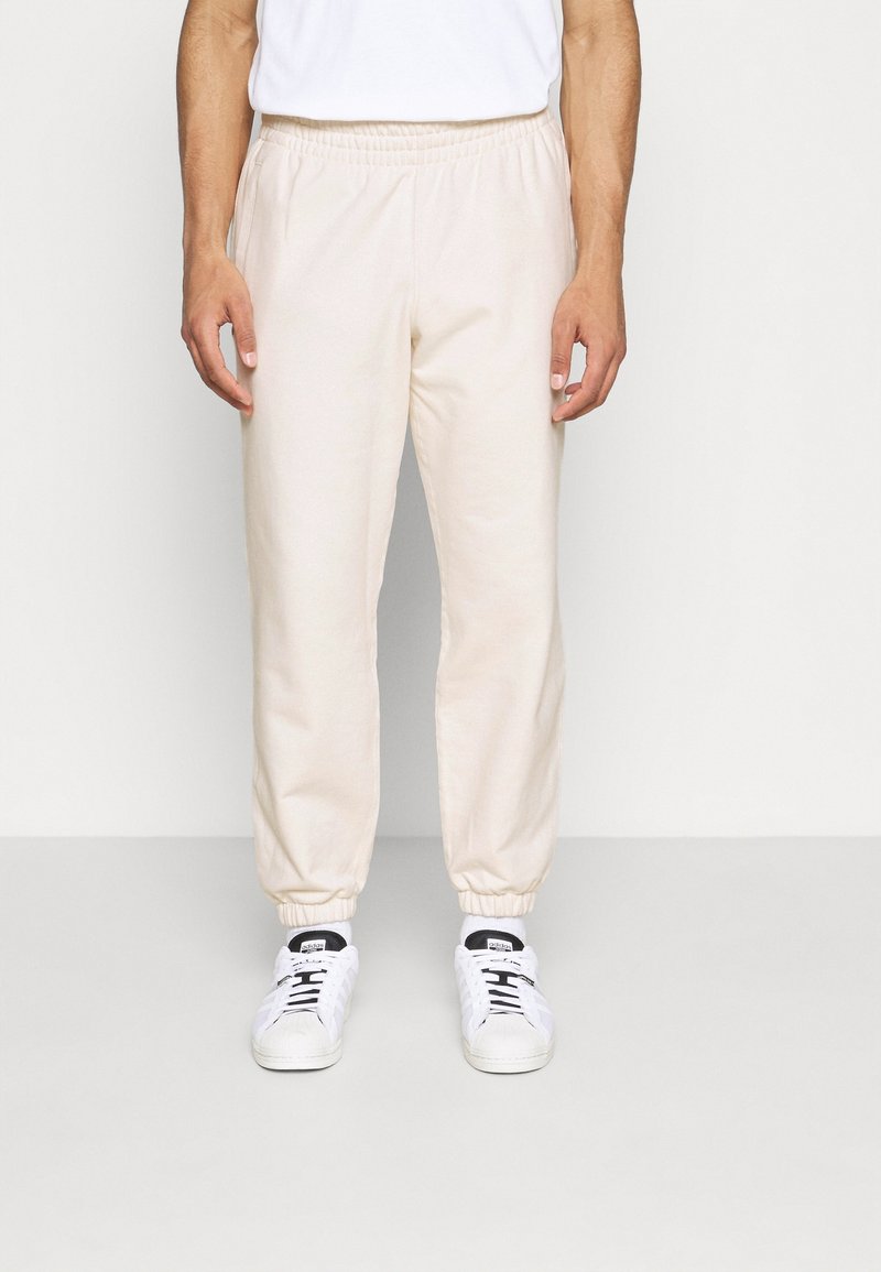 Man som bär krämfärgade joggingsbyxor, vita sneakers och en vit t-shirt som står mot en enkel ljus bakgrund.