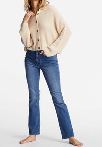 Beige ribbstickad kofta med krage och stora knappar, kombinerad med blå jeans med raka ben. Modellen står barfota.