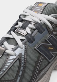 Γκρι και ασημί αθλητικό παπούτσι New Balance με λευκά κορδόνια, πλεκτό ύφασμα και τη σήμανση "NLOCK" στο πλάι και στη γλώσσα.