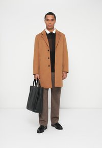 Cappotto lungo marrone in lana con rever a taglio, maglione nero in maglia, pantaloni a quadri marroni e una borsa a tracolla nera in pelle. Scarpe nere completano l'outfit.