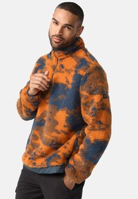 Fleece pulóver narancssárga és kék álcázó mintával, cipzáras gallérral és elasztikus derékkal. Puha tapintás, kontrasztos színek és laza fazon.