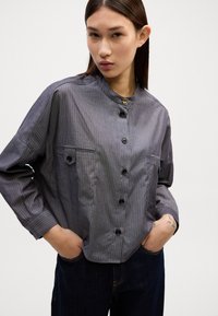 Een overhemd met knopen in een grijs en blauw gestreept patroon, met een Mandarin-kraag, twee voorzakken en zwarte knopen. Zachte stoftextuur.