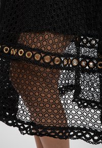 Robe en dentelle noire avec un motif circulaire, ourlet festonné et accents en œillets dorés le long d'une ceinture en tissu.