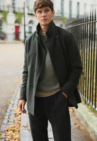 Tmavozelený vlnený kabát s veľkými gombíkmi, nosený cez sveter s rybej kosťou. Textúrované čierne nohavice a betónové pozadie.