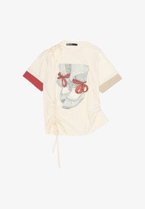 Chemise à manches courtes couleur crème avec des poignets de manche rouges et beiges, des liens froncés sur les côtés, et un motif représentant des pieds chaussés de ballerines avec des rubans rouges.