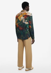 Camicia con motivo floreale in tonalità scure con fiori vivaci, abbinata a pantaloni beige larghi e calzature chiare. Mostrata vista posteriore.