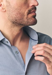 Kék kockás ing gombos gallérral, finom textúrával és szürke gombokkal, részben kigombolva a nyaknál. Bőrszín is látható.