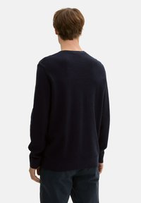 Dunkelblaues, geripptes Strickpullover mit langen Ärmeln, Rundhalsausschnitt und strukturiertem Stoff. Etikett auf dem linken Ärmel sichtbar.