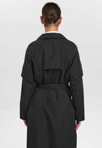 Svart trench coat med bälte, med en ryggslits, långa ärmar och knäppta manschetter. Slätt, lättviktigt tyg med en enkel design.