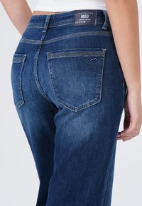 Close-up van een persoon die donkerblauwe spijkerbroek draagt met een label waarop staat "betoverende MOD denimcultuur" op de tailleband.