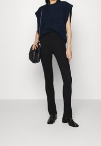 Navy gebreide oversized trui met korte mouwen, gecombineerd met zwarte geribde broek met zilveren stippen. Model houdt een zwarte gewatteerde tas vast.