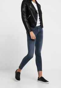 Svart lädermotorcykeljacka med kviltat axeldetaljer, vit t-shirt, mörkblå smala jeans med siddragkedjor, svarta sneakers.