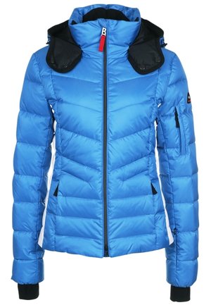 Personne portant une veste matelassée bleu vif avec capuche, pantalon noir et chaussures de randonnée noires, debout devant un fond uni.