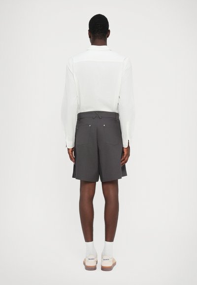 Shorts gris avec une coupe ajustée et des poches arrière, portés avec une chemise blanche à manches longues. Le mannequin se tient de dos sur un fond neutre.