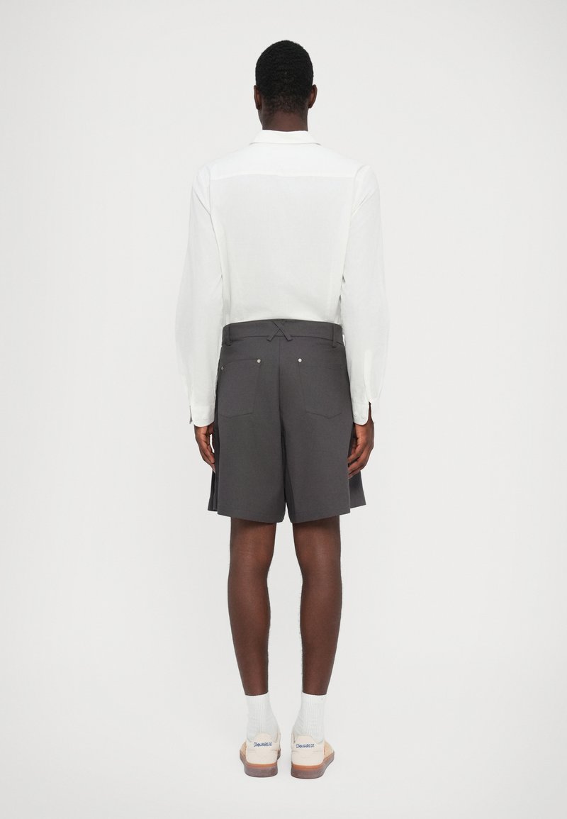 Grijze shorts met een getailleerde pasvorm en achterzakken, gedragen met een witte blouse met lange mouwen. Het model staat met de rug naar de camera op een neutrale achtergrond.