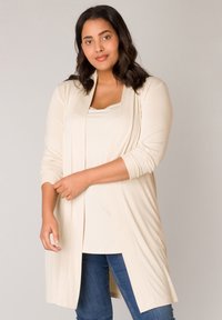 Beige lange cardigan met een open voorkant, lichtgewicht materiaal, lange mouwen en een relaxed fit, gedragen over een bijpassend topje en blauwe jeans.