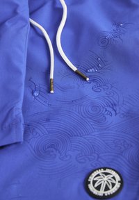 Veste bleue avec motif en relief ondulé, cordons de serrage blancs réglables avec embouts en métal, et un patch rond noir avec logo de palmier.