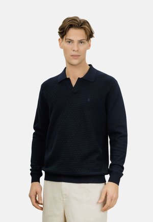 Ung mann med lyst brunt hår iført en marineblå teksturert langermet polotrøye og beige bukser, stående foran en ensfarget bakgrunn.