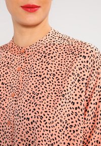 Femme portant un blouse pêche avec des taches léopard noires, boutonnée jusqu'au col, lèvres peintes avec un rouge à lèvres rouge vif.