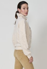 Sweatshirt beige avec un col montant, orné de broderies florales sur les manches et d'un cordon de serrage. Tissu doux et texturé avec des poignets côtelés.