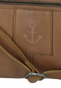 Borsa in pelle marrone con chiusura a zip, caratterizzata da un design con ancora in rilievo e la parola "HARBOUR". Include un dettaglio in metallo.